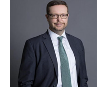 Male with brown hair and glasses wearing a suit smiling at camera