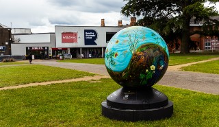 An image of a large decorative globe standing on some grass, with buildings and trees in the background