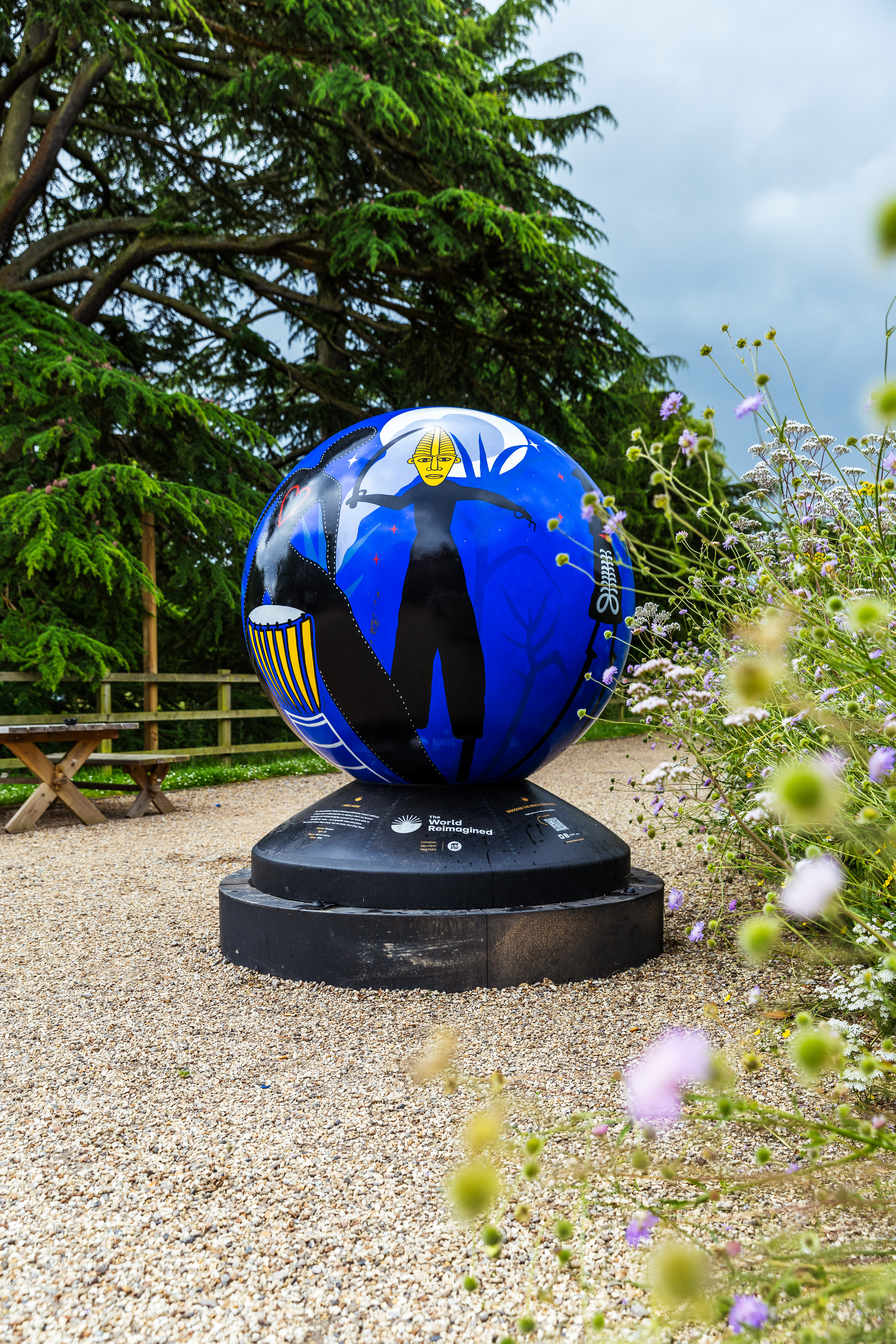 An image of a large decorative globe on some gravel, surrounded by flowers and trees