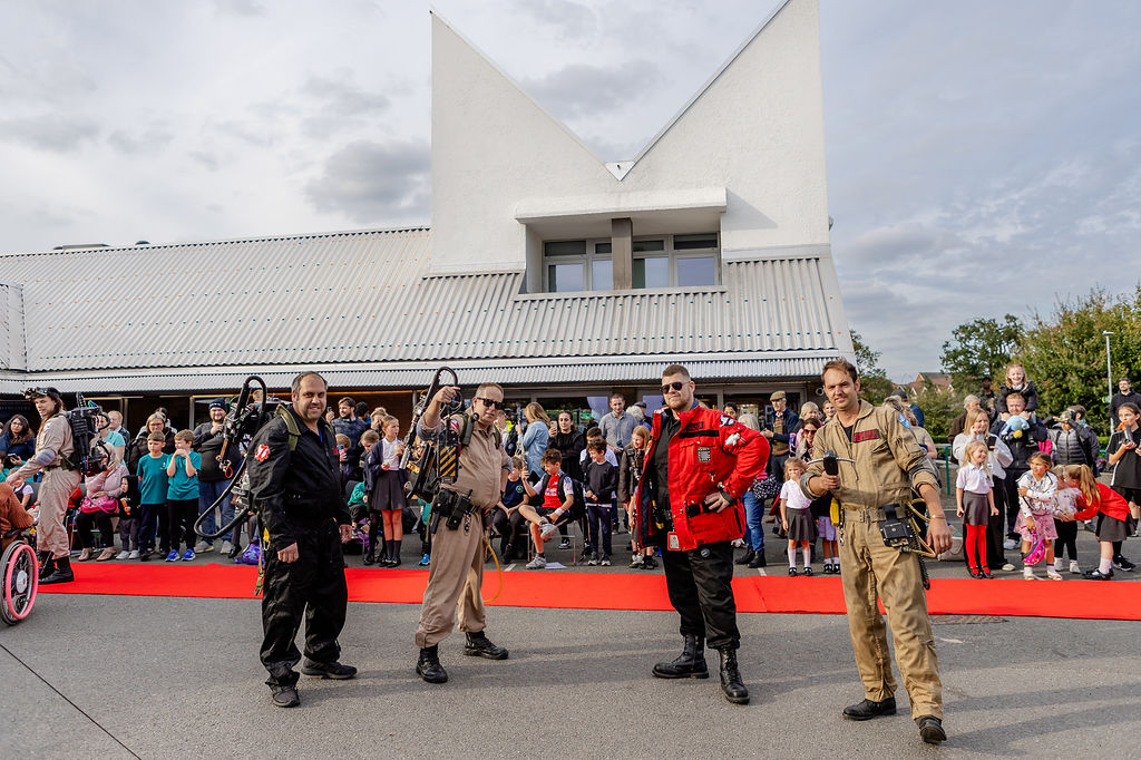 Four men dressed as characters from Ghostbusters, in front of a crowd of people, a red carpet and a community centre