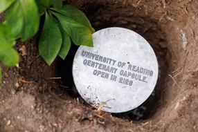 The top of a metal cylindrical capsule sitting in a hole in the ground. Engraved text reads 'University of Reading centenary capsule. Open in 2126'