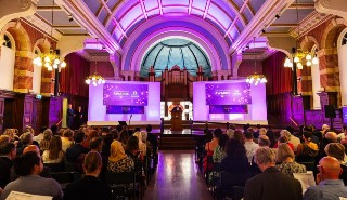A large hall with a stage and audience and purple lighting