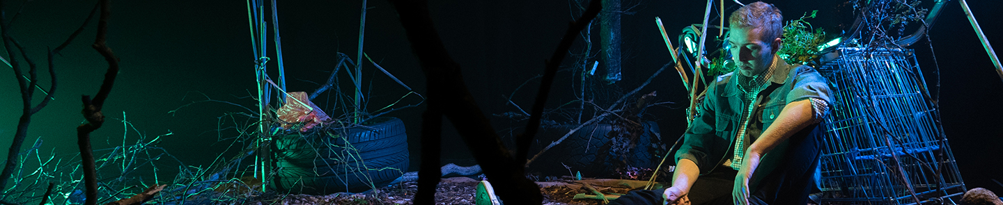 An actor sits pensively on a stage dressed to look like littered scrubland