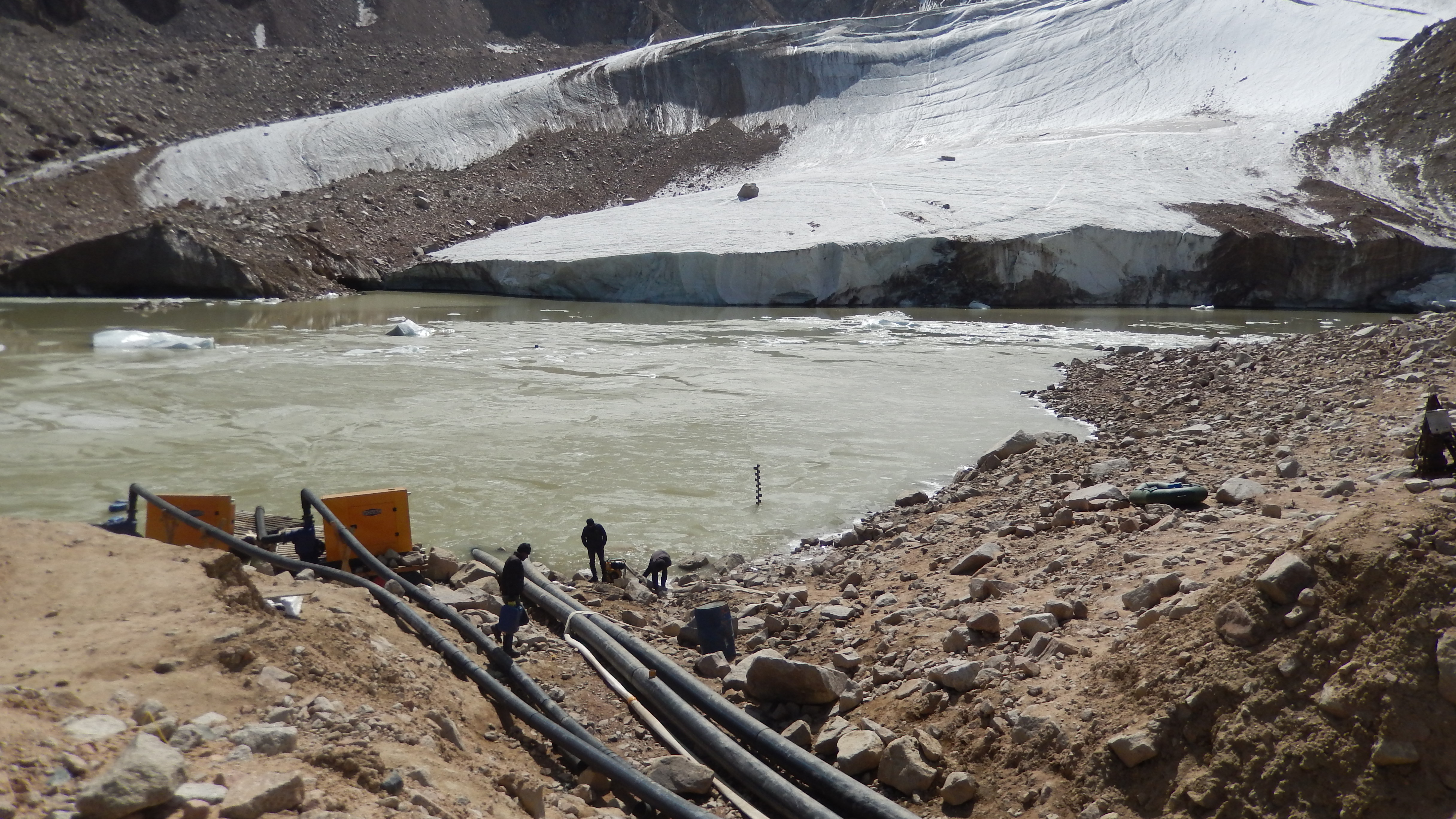 Men inspecting large pipes going into a lake in a rocky, glacial landscape.